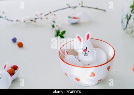 Des récipients amusants pour oeufs de Pâques remplis d'œufs de bonbons et de chocolat avec des oreilles de lapin dans un bol sur la table blanche de cuisine. Pâques festives du printemps Banque D'Images