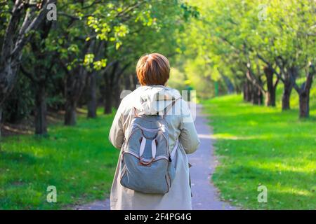 Une femme aux cheveux rouges dans un trench-coat beige avec un sac à dos rayé randonnée dans un parc de printemps. Le visage est méconnaissable, une vue de l'arrière. Un enroulement Banque D'Images
