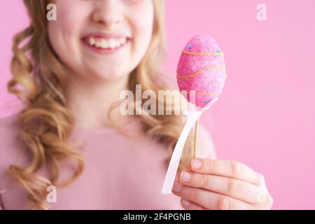 Gros plan sur une fille moderne heureuse avec de longs cheveux blond ondulés avec un œuf de pâques isolé sur fond rose. Banque D'Images