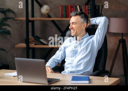 Homme d'affaires décontracté et gai, directeur, homme de race blanche, assis dans le bureau, habillé de style, repose du travail, jetant sa main derrière sa tête, rêves de vacances, regarde loin, sourires Banque D'Images