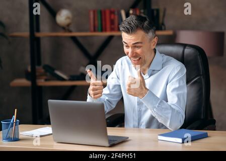 Homme d'affaires heureux, responsable, qui a réussi et qui a confiance en lui, communique par conférence téléphonique, en ligne avec ses collègues, montre un geste d'approbation, des gestes avec ses mains, se réjouit de la réussite, sourit Banque D'Images