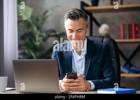 Chat en ligne. Un homme d'affaires caucasien joyeux et charmant est assis à son bureau, à l'aide d'un téléphone mobile pendant son travail, en naviguant sur Internet, en répondant à un e-mail et en envoyant un SMS à un ami ou à une famille, en souriant Banque D'Images
