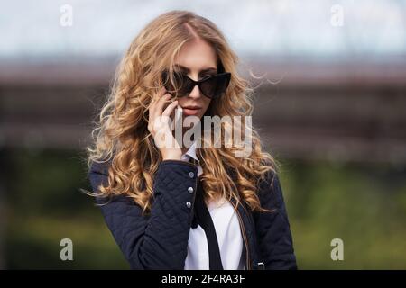 Jeune femme d'affaires en lunettes de soleil appelant sur téléphone portable élégant modèle tendance en veste bleu foncé et chemise blanche Banque D'Images