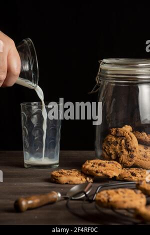 Vue de la main de la femme versant du lait dans un verre, sur une table en bois avec des biscuits au chocolat, fond noir, vertical Banque D'Images