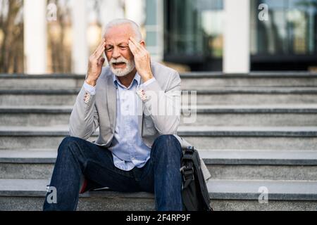 L'homme d'affaires senior est fatigué du travail. Il est assis devant le bâtiment de l'entreprise et a des maux de tête. Banque D'Images