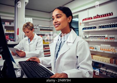 Jeune femme souriante travaillant en pharmacie à l'aide d'un ordinateur avec un senior collègue utilisant une tablette numérique dans un pharmacien derrière le comptoir Banque D'Images