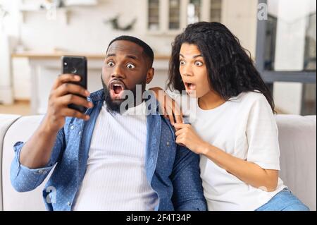 Se réveille en vie. Surpris couple multiracial regarde l'écran de smartphone, ils ont la bouche ouverte avec étonnement, hurlant wow, a été incroyable offre ou des nouvelles Banque D'Images