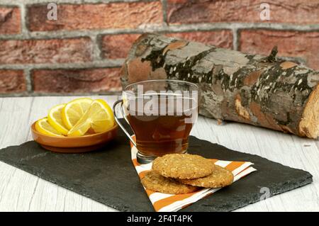 Tasse de thé, tranches de citron et biscuits aux flocons d'avoine, sur la table au-dessus de l'assiette en pierre, décoration de bois en rondins avec fond de mur en brique, concept de nourriture saine Banque D'Images