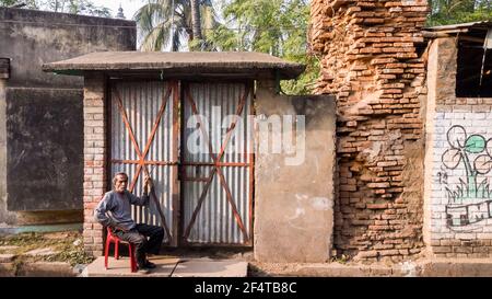 Murshidabad, Bengale-Occidental, Inde - janvier 2018: Un homme assis devant les volets fermés d'un magasin de grungy dans les rues de la ville de Murshidabad. Banque D'Images