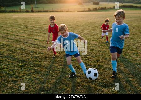 Enfants jouant au football amateur. Des garçons gais vêtues de robes de football ayant du plaisir à chasser le ballon à l'extérieur sur le terrain au coucher du soleil. Banque D'Images