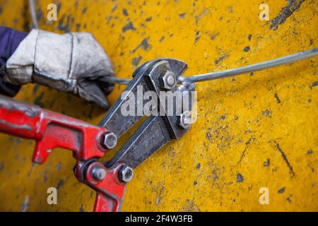 Les grosses pinces coupantes coupantes coupantes la tige en fil d'aluminium. Fond de sol peint en béton jaune. Main de l'employé dans des gants de protection en coton. Gros plan. Banque D'Images