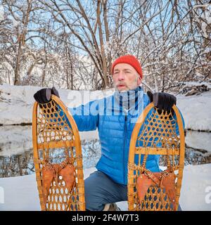 Homme âgé avec des raquettes en bois classiques (Bear Paw) sur une rive de ruisseau, paysage d'hiver au Colorado Banque D'Images