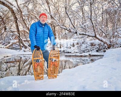Homme âgé avec des raquettes en bois classiques (Bear Paw) sur une rive de ruisseau, paysage d'hiver au Colorado Banque D'Images