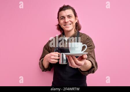 Un serveur masculin offrant une tasse de café isolée sur fond rose, un homme souriant dans un tablier donne une tasse de boisson à l'appareil photo. Concentrez-vous sur la tasse Banque D'Images