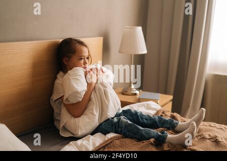 Énervé petite fille pleurant assis seul en position foetale se cachant le visage et se balançant sur le lit embrassant grand oreiller. Banque D'Images