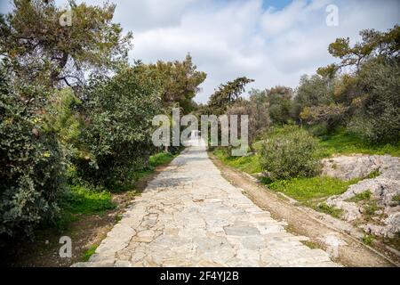 Colline de Filopappou, Athènes, Grèce. Chemin pavé sous l'ancienne roche grecque de l'Acropole, entre les arbres de la forêt de Philopapos. Jour, ciel bleu ciel nuageux, Banque D'Images