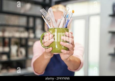 Les choses nécessaires. Gros plan des mains de la femme tenant des outils, des brosses pour créer un bol en céramique d'argile fait à la main dans un atelier de poterie Banque D'Images