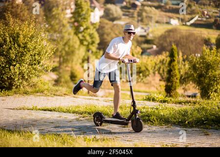 Joyeux et décontracté homme dans le chapeau et les lunettes de soleil équitation électrique scooter dans le parc d'été de la ville Banque D'Images