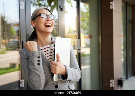 Prêt à travailler. Belle et heureuse femme d'affaires mûre tenant un ordinateur portable et souriant tout en se tenant près du bâtiment de bureau Banque D'Images