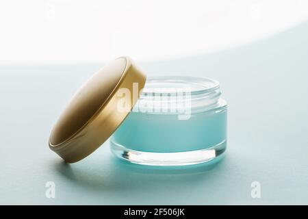 Gel hydratant pour le visage dans un pot ouvert avec couvercle doré sur fond bleu clair. Contenant en verre de crème hydratante pour le visage. Beauté, soins de la peau. Banque D'Images