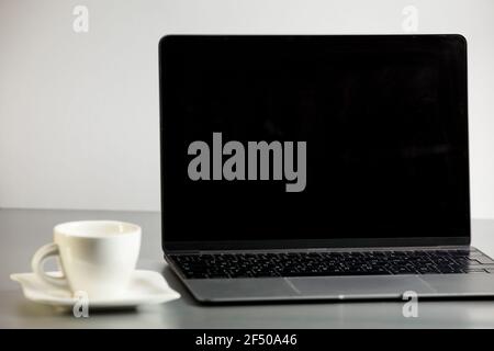 Ordinateur portable moderne ouvert et tasse à café sur soucoupe sur table grise sur fond blanc. Travail à distance, bureau à domicile. Moke up avec un ordinateur portable et une tasse de café. Banque D'Images