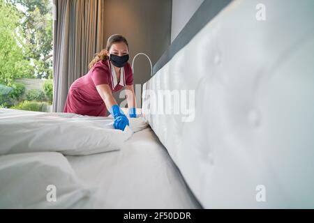 Femme de ménage dans un masque facial faisant le lit à l'hôtel chambre Banque D'Images