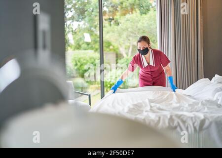 Femme de ménage dans un masque facial faisant le lit à l'hôtel chambre Banque D'Images