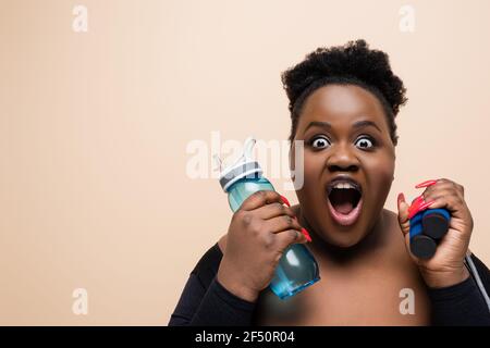 une femme surdimensionnée afro-américaine choquée tenant une bouteille de sport et sautant corde isolée sur beige Banque D'Images