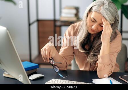 Fatigué bouleversé mature gris-cheveux asiatique, femme d'affaires, gestionnaire ou professeur, prend une pause de travail en ligne, massage sa tête avec sa main, a un mal de tête, s'assoit à la table, a pris des lunettes, besoin de repos Banque D'Images
