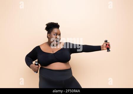 femme afro-américaine de grande taille excitée dans les vêtements de sport tenant des haltères isolé sur beige Banque D'Images