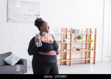 joyeuse femme afro-américaine en surpoids dans les vêtements de sport tenant une bouteille de sport à la maison Banque D'Images