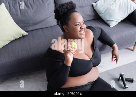 femme afro-américaine de grande taille et enjouée dans des vêtements de sport tenant le vert apple Banque D'Images