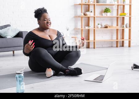 femme afro-américaine souriante et de taille plus regardant un entraînement en ligne sur un ordinateur portable assis dans une posture de yoga sur un tapis de fitness Banque D'Images