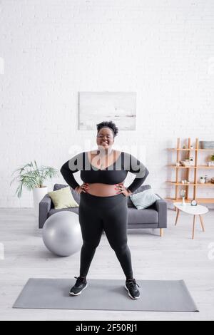 femme afro-américaine de grande taille dans les vêtements de sport debout, les mains sur les hanches, sur le tapis de fitness Banque D'Images