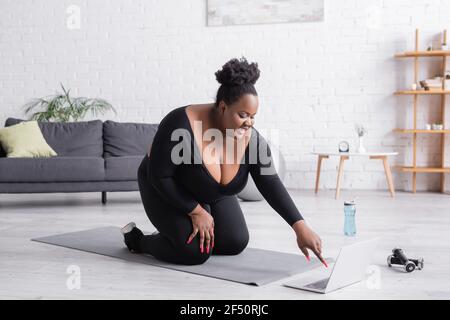 bonne femme afro-américaine de taille plus regardant l'entraînement sportif sur un ordinateur portable assis sur un tapis de fitness dans la salle de séjour Banque D'Images