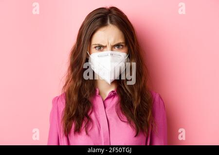 Covid-19, concept de quarantaine et de distanciation sociale. Gros plan portrait d'une femme en colère dans un appareil respiratoire frogné, qui a l'air contrarié et judgmental, rose Banque D'Images