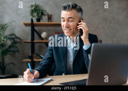 Manager ou chef d'entreprise expérimenté, intelligent et confiant, au caucase, assis au bureau, dans un costume élégant, communique avec un client par smartphone, prend des notes, sourit Banque D'Images