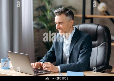 Un homme d'affaires ou un responsable d'un costume classique, connu pour son succès, utilise un ordinateur portable, communique via la visioconférence, les réunions en ligne, se trouve au bureau, saisit sur un clavier, sourit Banque D'Images