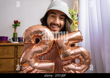 Beau jeune homme latino-asiatique tenant une montgolfière métallique, célébrant son 25e anniversaire. Ballon métallique avec le numéro 25. Banque D'Images