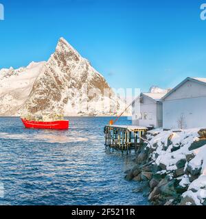 Maisons traditionnelles norvégiennes en bois sur la rive de Reinefjorden et bateaux de pêche avec des montagnes enneigées en arrière-plan . Emplacement: Île de Toppoya, Banque D'Images