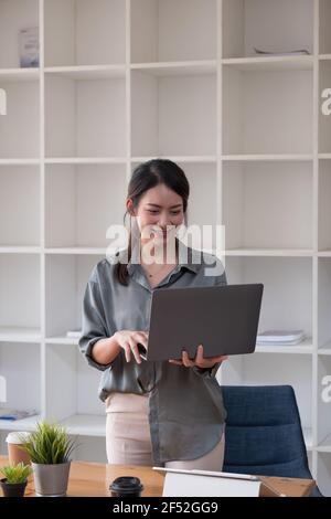 Jeune femme asiatique debout et tenant un ordinateur portable de navigation sur Internet au bureau à la maison. Banque D'Images