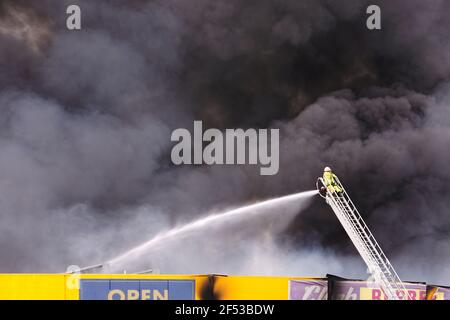 Pompier en action sur une échelle pulvérisant de l'eau sur un bâtiment au feu avec d'énormes nuages de fumée noire remplissant le ciel. Banque D'Images