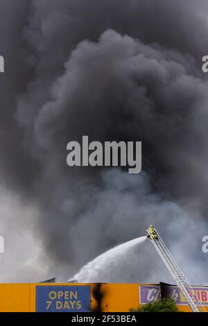 Pompier en action sur une échelle pulvérisant de l'eau sur un bâtiment au feu avec d'énormes nuages de fumée noire remplissant le ciel. Banque D'Images