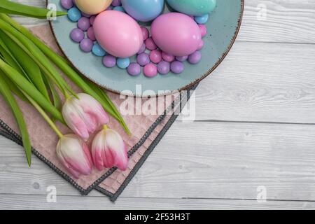 Œufs de Pâques pastel et bonbons au chocolat pastel dans une assiette bleue et tulipes blanches roses, sur fond de bois blanc. Banque D'Images