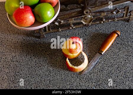 Une pomme à moitié pelée et un petit couteau sur un banc de cuisine avec un ensemble vintage de balances et de pommes rouges et vertes. Banque D'Images