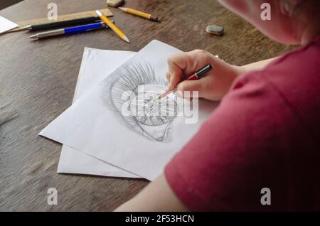 Une femme d'âge moyen tire avec un crayon sur du papier blanc. Une femme adulte s'assoit à une table en bois et dessine un œil humain. Art, loisirs et loisirs. Artis Banque D'Images