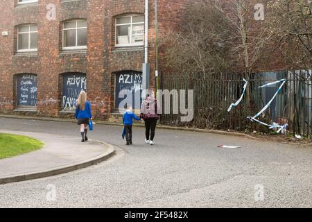 Stockton-onTees, Royaume-Uni. 24 mars 2021. Une femme et deux jeunes enfants en uniforme scolaire marchent devant un trou dans une clôture couverte de bande de police tandis que la police de Cleveland recherche l'homme manquant Andrew Stones près de Dunmail Road, Stockton. Credit Jason Brown / Alamy Live News Banque D'Images