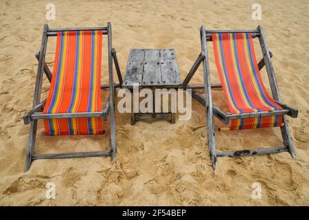 Deux chaises longues en toile colorées et vides et table en bois sur la plage de sable de la station balnéaire en Thaïlande, vue avant, grand angle Banque D'Images