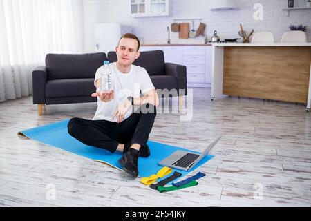 L'homme entre pour le sport à la maison. L'athlète boit de l'eau tout en étant assis sur le sol à la maison, un mode de vie sain Banque D'Images