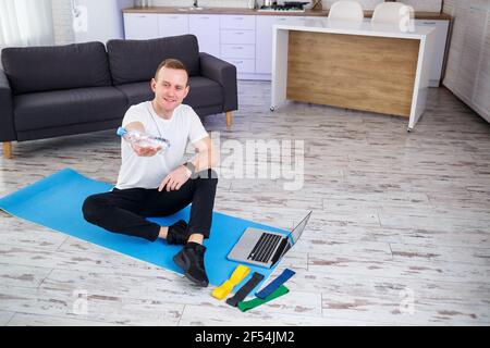L'homme entre pour le sport à la maison. L'athlète boit de l'eau tout en étant assis sur le sol à la maison, un mode de vie sain Banque D'Images